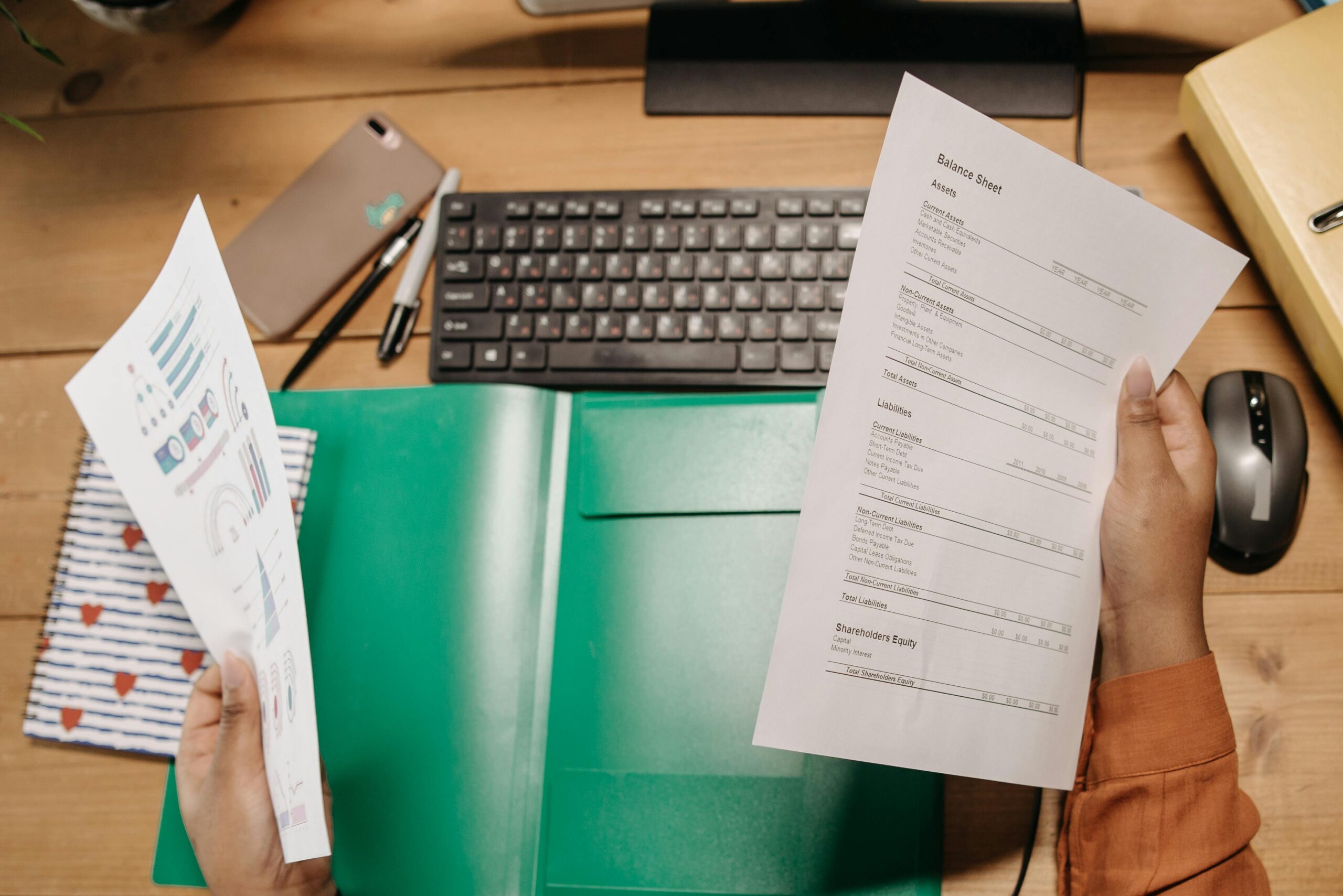 Excelentnosť dokumentácie Hands holding and reviewing balance sheets over a desk with a keyboard and stationery.